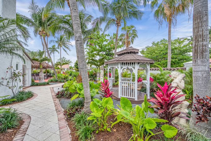 Lush tropical courtyard with tall palm trees and a white wooden gazebo surrounded by bright pink and green foliage, stone statues and a curved paved walkway under a blue sky.