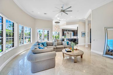 Spacious waterfront open‑concept living room with curved bay windows, light gray sectional sofa, round rustic coffee table, marble floors, modern ceiling fan, and canal views with palm trees.