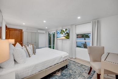 Bright modern coastal-style bedroom with king bed in white linens, decorative pillows, wall-mounted TV showing a palm beach scene, sliding glass door and window with curtains, area rug and small desk with chair
