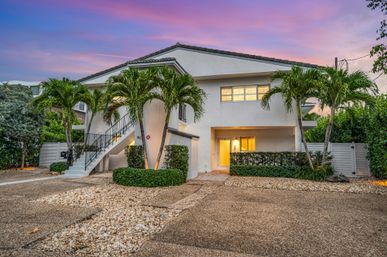 Two-story white coastal home at dusk with exterior staircase, glowing entryway, palm trees and pebble driveway beneath a pink-purple sunset sky