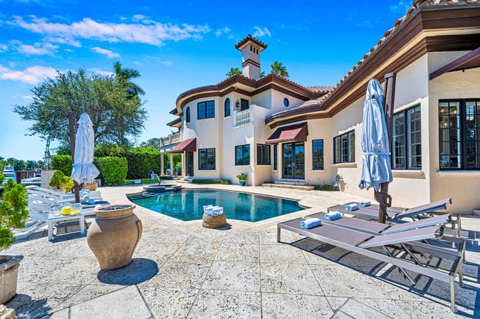 Sunny waterfront Mediterranean-style villa with private swimming pool, stone patio, lounge chairs with rolled towels, closed umbrellas, terracotta planters and a boat dock under a bright blue sky.