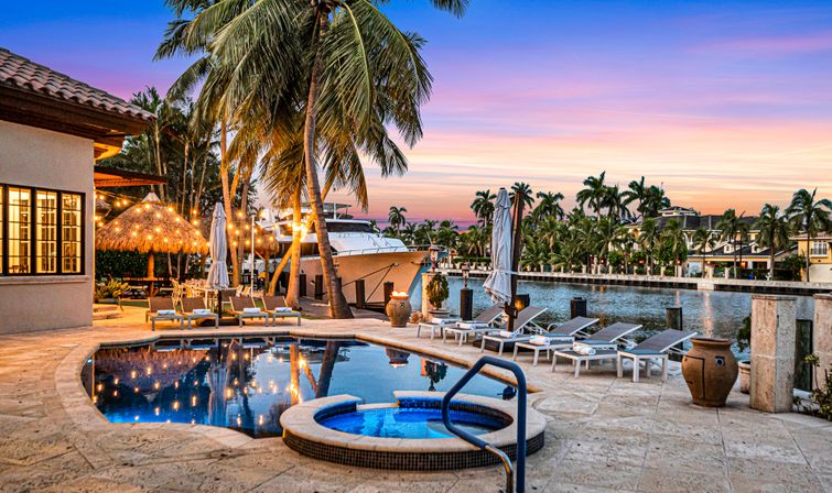 Sunset at a luxury waterfront villa with a tiled pool and round hot tub in the foreground, sun loungers and string-lit tiki patio, palm trees and a docked yacht along a calm marina.
