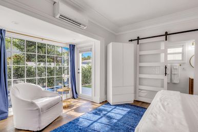 Sunlit coastal-style bedroom with large grid window and patio door to a garden, white slipcovered armchair, blue area rug, sliding barn door, white wardrobe and wall-mounted air conditioner.