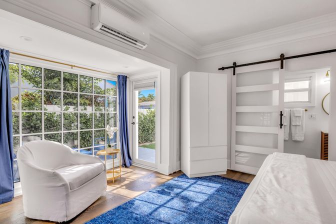 Sunlit coastal-style bedroom with large grid window and patio door to a garden, white slipcovered armchair, blue area rug, sliding barn door, white wardrobe and wall-mounted air conditioner.