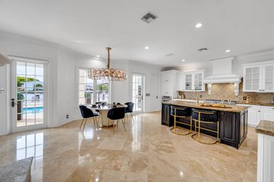 Bright open-concept luxury kitchen and dining area with polished marble floors, dark island with gold-accent stools, round dining table under a sparkling chandelier, white cabinetry, and French doors opening to a pool and waterfront view.