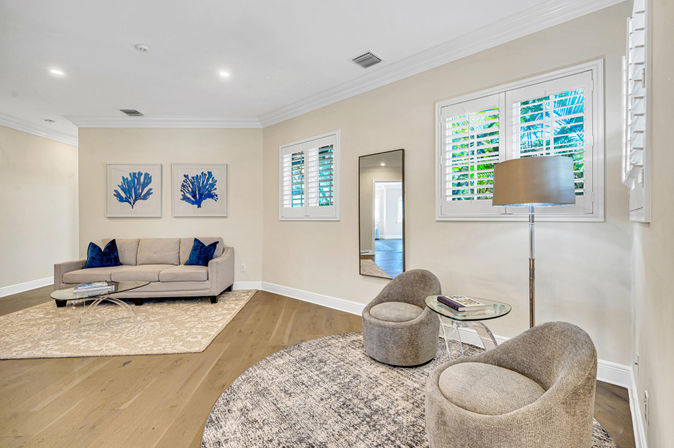 Bright modern living room with a beige sofa and blue coral wall art, two gray swivel chairs on a round rug, glass side table and floor lamp, plantation shutters on windows and light oak hardwood floors in a neutral, coastal-inspired setting.