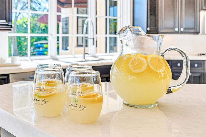 Sunlit modern kitchen island with a glass pitcher of fresh lemonade garnished with lemon slices and three filled glasses on a marble countertop, ready to serve.