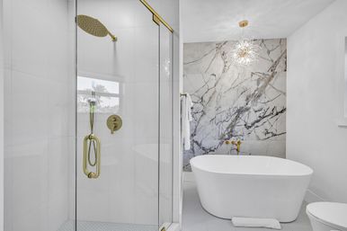 Spa-like modern bathroom with freestanding white soaking tub, glass-enclosed shower with brass fixtures, dramatic marble accent wall and starburst pendant light.