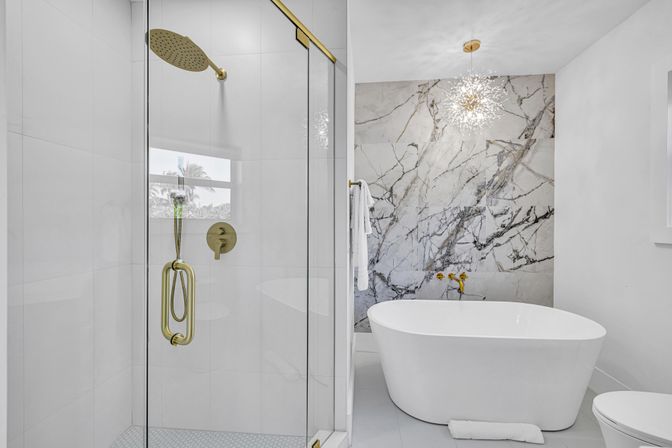 Spa-like modern bathroom with freestanding white soaking tub, glass-enclosed shower with brass fixtures, dramatic marble accent wall and starburst pendant light.