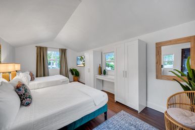 Bright, airy coastal bedroom with two white-made beds, wood floors, white wardrobes and desk, rattan chair, potted plants and palm-tree view through the windows.