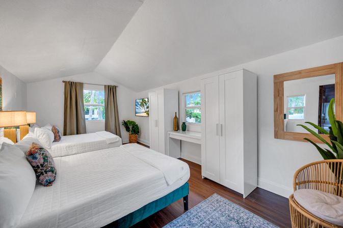 Bright, airy coastal bedroom with two white-made beds, wood floors, white wardrobes and desk, rattan chair, potted plants and palm-tree view through the windows.