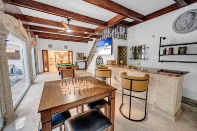 Sunny Mediterranean-style game room with beamed ceiling, chess table in foreground, pool table, stone bar with yellow stools, TV and sliding patio doors.