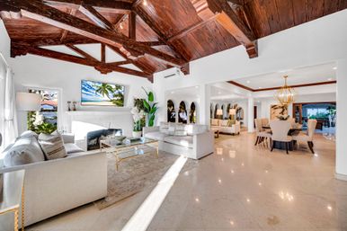 Sunlit open-concept living and dining room with dark exposed wooden-beam ceiling, marble floors, neutral sofas around a fireplace, glass coffee table, tropical wall art, arched windows and elegant dining table.