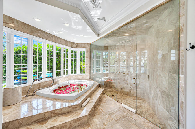 Sunlit luxury marble bathroom with glass-enclosed shower and sunken soaking tub strewn with rose petals, wall of windows overlooking a lush garden and pool