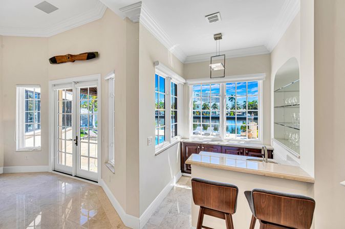 Sunlit waterfront kitchen nook with marble floors, breakfast bar and two wooden stools, bay windows and French doors framing a canal view with palm trees, and a vintage wooden propeller mounted above the door.