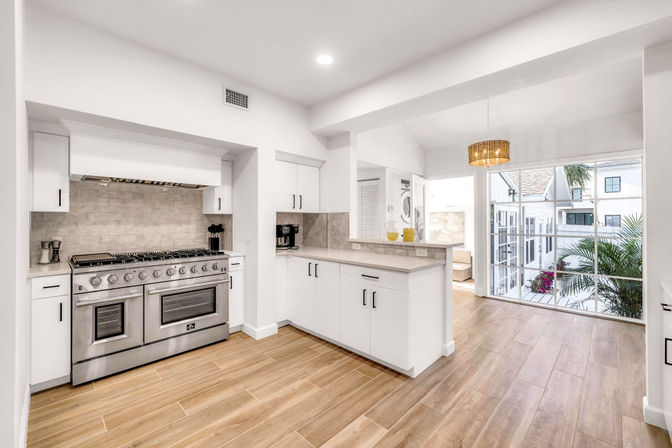 Bright, airy open-plan kitchen with a stainless-steel professional range, white cabinetry, wood-look tile flooring, breakfast bar, and sunlit sliding doors opening to a balcony with tropical plants