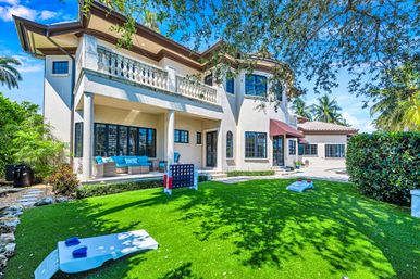 Sunlit Mediterranean-style two-story home with covered patio, turquoise outdoor seating, palm-lined backyard and lawn games (cornhole and giant Connect Four) on a manicured green lawn.