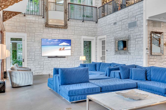 Coastal-inspired modern living room with large royal-blue modular sectional, stone accent wall, wall-mounted TV showing a beach scene, high ceilings with upstairs balcony railings, glass doors and contemporary coffee table.