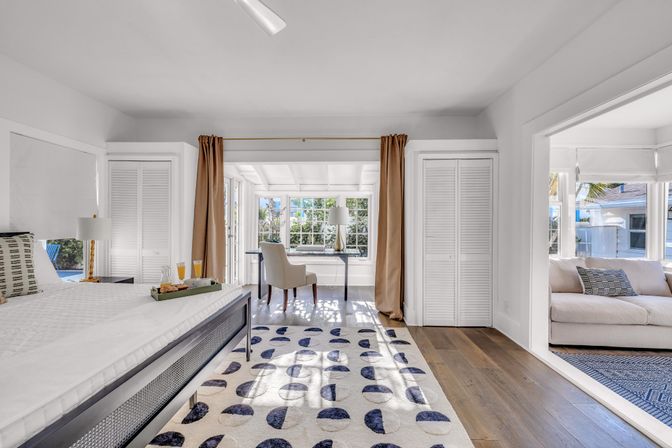 Sunlit modern coastal-style bedroom with low platform bed, blue-and-white patterned rug, hardwood floors, beige curtains framing a bright windowed desk nook, and an adjacent sunroom with a light sofa.