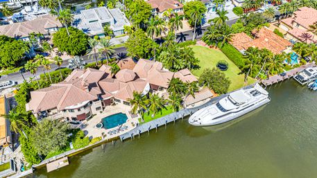 Aerial drone view of a luxury Mediterranean-style waterfront mansion with red tile roofs, palm trees, curved swimming pool, private dock and a large white yacht moored on a calm canal