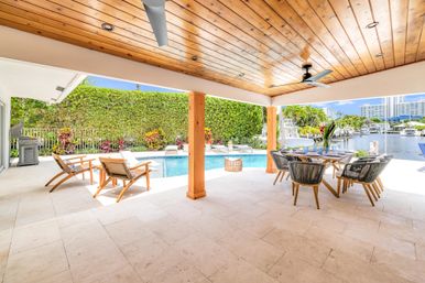 Sunny covered waterfront patio with wood-paneled ceiling and fans, poolside lounge chairs, outdoor dining set and view of a pool and canal with boats.