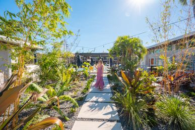 Sun-drenched, palm-lined boutique hotel courtyard with lush tropical plants flanking a stepping-stone path; a woman in a pink sundress strolls toward a pool area under string lights and a bright blue sky.