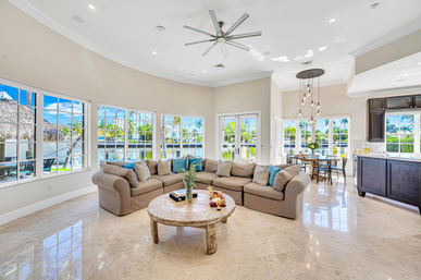 Sunlit waterfront open-concept living room with large curved windows overlooking a canal and palm trees, neutral L-shaped sectional with blue accent pillows, round rustic coffee table, glossy marble floors, dining area and kitchen in view — relaxed coastal vibe.