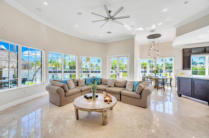 Sunlit waterfront open-concept living room with large curved windows overlooking a canal and palm trees, neutral L-shaped sectional with blue accent pillows, round rustic coffee table, glossy marble floors, dining area and kitchen in view — relaxed coastal vibe.