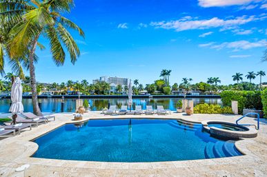 Waterfront tropical pool and spa with palm trees, sun loungers and umbrellas overlooking a calm canal under a bright blue sky.