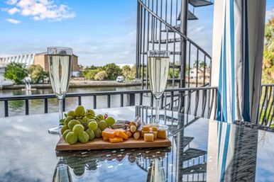 Two flutes of sparkling wine and a cheese-and-grape charcuterie board on a glass table on a waterfront balcony with canal view and spiral staircase