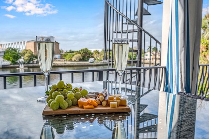 Two flutes of sparkling wine and a cheese-and-grape charcuterie board on a glass table on a waterfront balcony with canal view and spiral staircase