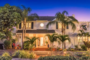 Twilight coastal-style two-story house with palm trees, warm porch lights, and lush tropical landscaping.