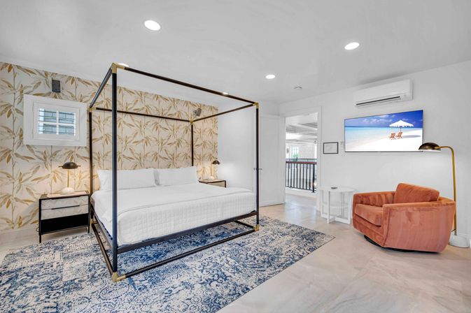 Bright beach-inspired bedroom with black metal four-poster king bed and white bedding, tropical leaf wallpaper accent wall, blue patterned area rug, coral velvet swivel armchair, bedside lamps, floor lamp, and wall-mounted TV showing a beach scene.
