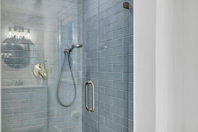 Modern glass-enclosed shower with coastal blue subway tile, brushed-nickel handheld showerhead and vertical pull handle, reflection of round vanity mirror and light fixture