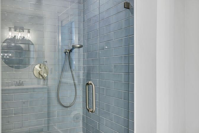 Modern glass-enclosed shower with coastal blue subway tile, brushed-nickel handheld showerhead and vertical pull handle, reflection of round vanity mirror and light fixture