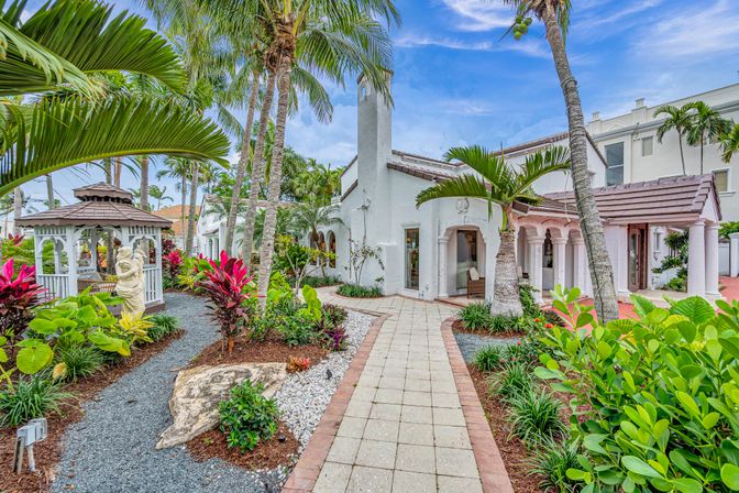 Tropical South Florida–style white stucco villa with tiled roof, palm-lined paved walkway winding through lush landscaped gardens to a white gazebo and arched entry.