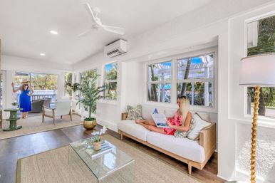 Sunlit coastal living room with white walls and hardwood floors, rattan sofa with a person in a pink floral dress reading a magazine, another person in a blue sundress and sunhat looking out large windows at palm trees and outdoor seating, glass coffee table, indoor plants and ceiling fan for a bright beachside bungalow vibe.