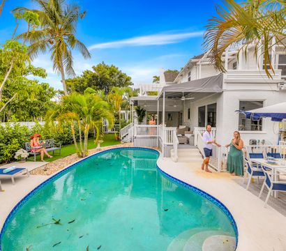 Sunny tropical backyard oasis with a kidney-shaped turquoise pool, white two-story coastal house and covered porch, outdoor dining under a blue umbrella, palm trees, lounge chairs, and people relaxing and socializing.