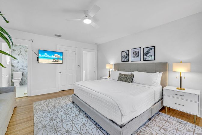 Bright modern bedroom with king bed and tufted gray headboard, white linens, patterned rug, hardwood floors, ceiling fan, gold bedside lamps, wall-mounted TV showing a beach and an en-suite marble-tiled bathroom.