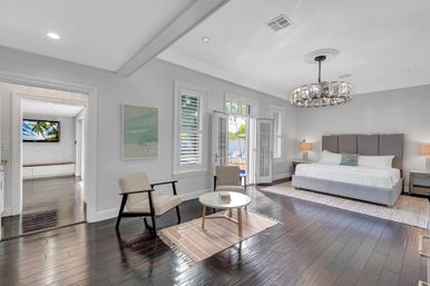 Bright modern master bedroom with king bed and gray upholstered headboard, matching nightstands and lamps, crystal chandelier, sitting area with two mid-century chairs and round coffee table on a rug, dark hardwood floors and French doors opening to a poolside patio.