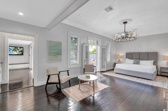Bright modern master bedroom with king bed and gray upholstered headboard, matching nightstands and lamps, crystal chandelier, sitting area with two mid-century chairs and round coffee table on a rug, dark hardwood floors and French doors opening to a poolside patio.