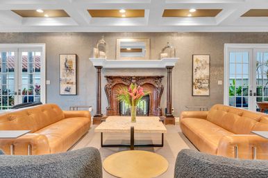Sunlit luxury living room with matching tan leather sofas facing an ornate carved wood fireplace, marble coffee table topped with tropical flowers, coffered ceiling with recessed lights and French doors opening to a patio.