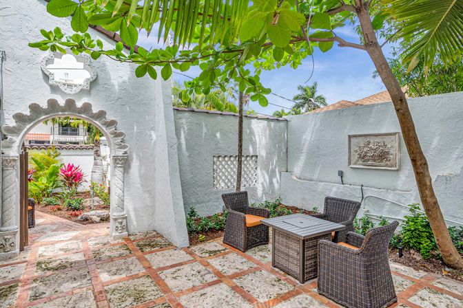 Cozy tropical stucco courtyard with ornate arched gateway, palm trees, wicker patio chairs with orange cushions around a square table on a stone-tile patio
