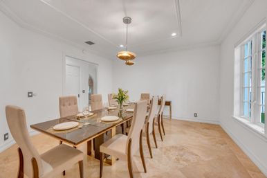 Sleek modern dining room with eight beige upholstered chairs around a glass-top table, marble floors, white walls, pendant light, large window, and a set table with plates and a floral centerpiece.