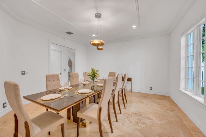 Sleek modern dining room with eight beige upholstered chairs around a glass-top table, marble floors, white walls, pendant light, large window, and a set table with plates and a floral centerpiece.