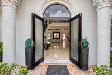 Open double black doors of a Mediterranean-style luxury home with arched transom, stucco columns and potted topiaries framing a polished marble foyer leading to an elegant living room and curved staircase.