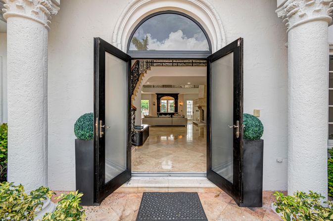 Open double black doors of a Mediterranean-style luxury home with arched transom, stucco columns and potted topiaries framing a polished marble foyer leading to an elegant living room and curved staircase.