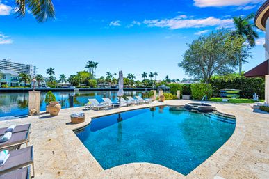 Sunny tropical waterfront backyard with a sparkling blue pool and hot tub, sun loungers on a travertine deck, palm trees and canal views