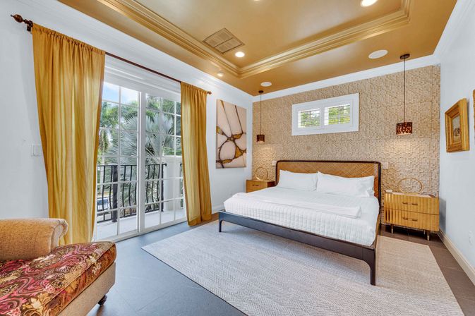 Sunny tropical master bedroom with king bed and woven headboard, textured accent wall, hanging pendant lights, gold tray ceiling, large glass doors with mustard curtains opening to a balcony with palm trees, patterned chaise and neutral area rug.