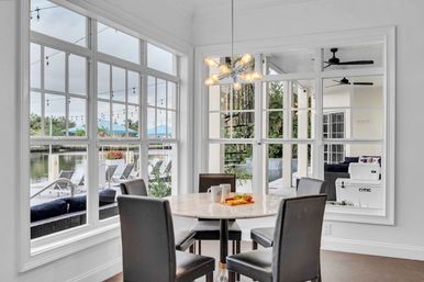 Airy waterfront dining nook with round marble table and four black chairs, large paned windows overlooking a dock, pool and string-lit patio